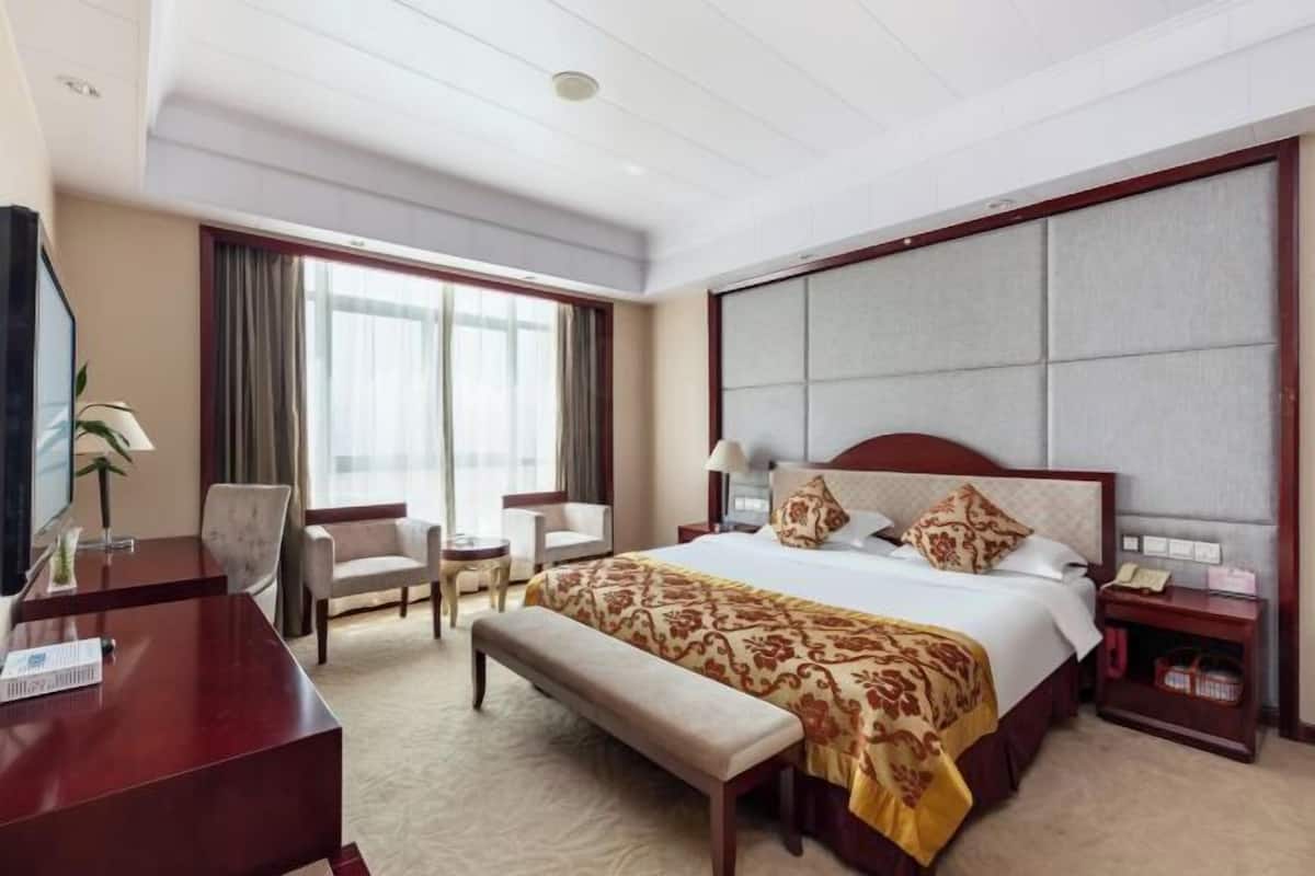 A hotel room with a large bed, a desk, a chair, a bench, and a television.