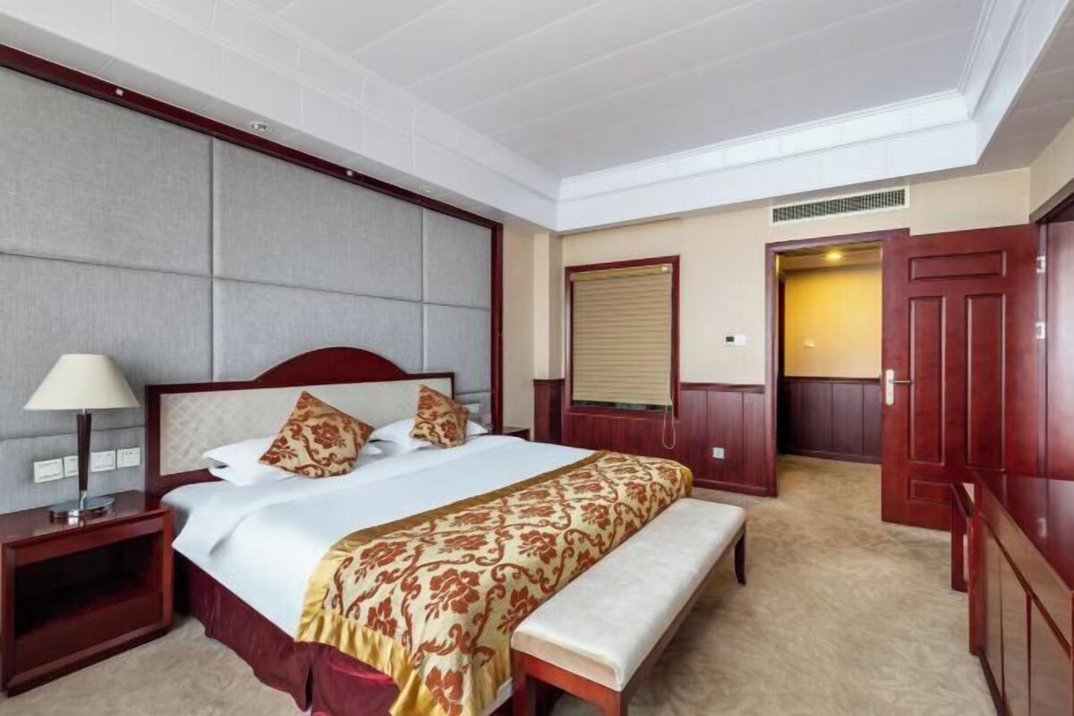 A hotel room with a large bed, a bedside table with a lamp, a bench, and a wardrobe.