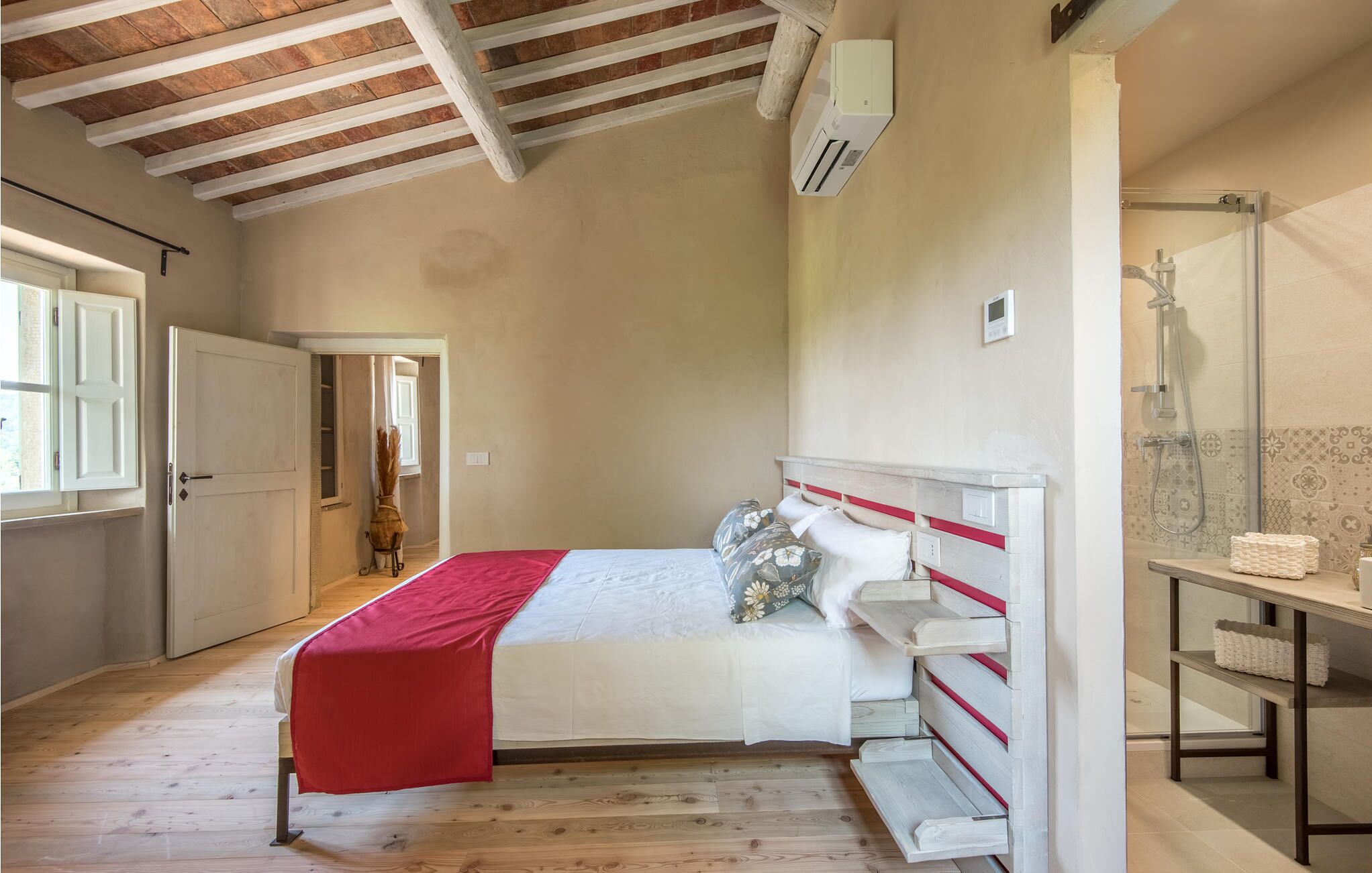 A bedroom with a wooden floor, a bed with a red blanket, a bedside table, and a bathroom with a shower.
