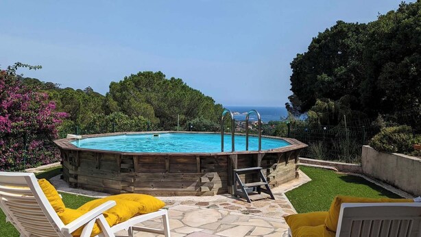 A pool with a wooden deck, two white lounge chairs with yellow cushions, and a view of the sea.