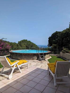 A pool with a wooden deck, two white lounge chairs with yellow cushions, and a view of the sea.