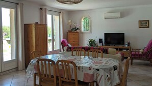 A dining area with a round table, chairs, a hanging light fixture, a television, and a window with curtains.