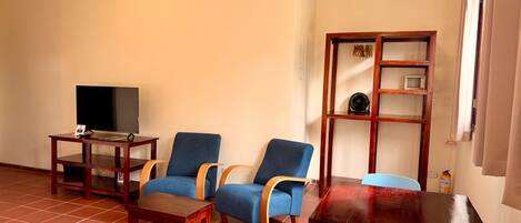 Una sala de estar con televisor, dos sillones azules, una mesa de centro de madera y una estantería de madera.