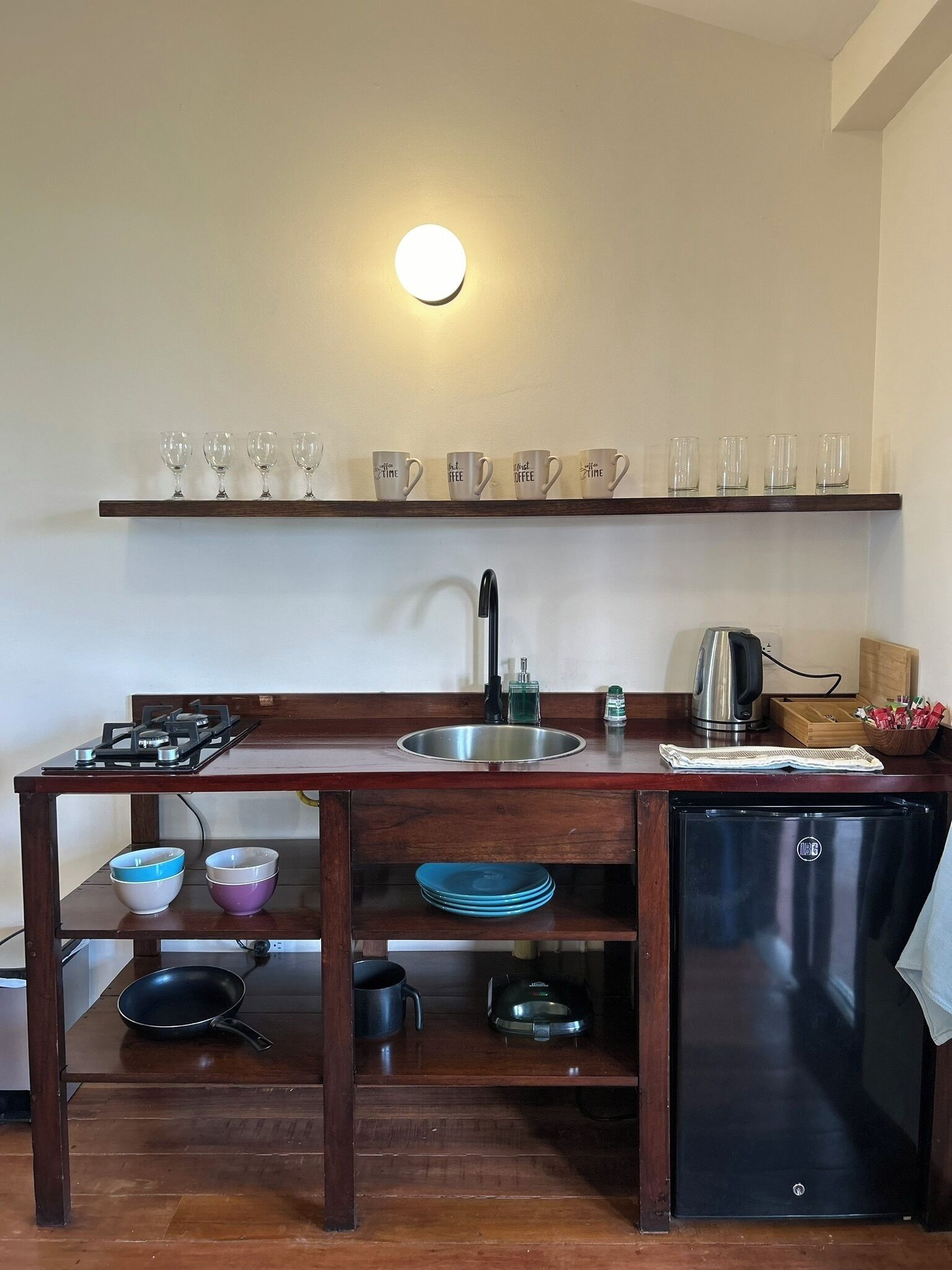 A compact kitchen with a gas stove, sink, and a black refrigerator. There are shelves above the sink holding glasses and cups, and a wooden table with bowls and a frying pan.