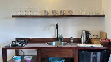 A compact kitchen with a gas stove, sink, and a black refrigerator. There are shelves above the sink holding glasses and cups, and a wooden table with bowls and a frying pan.