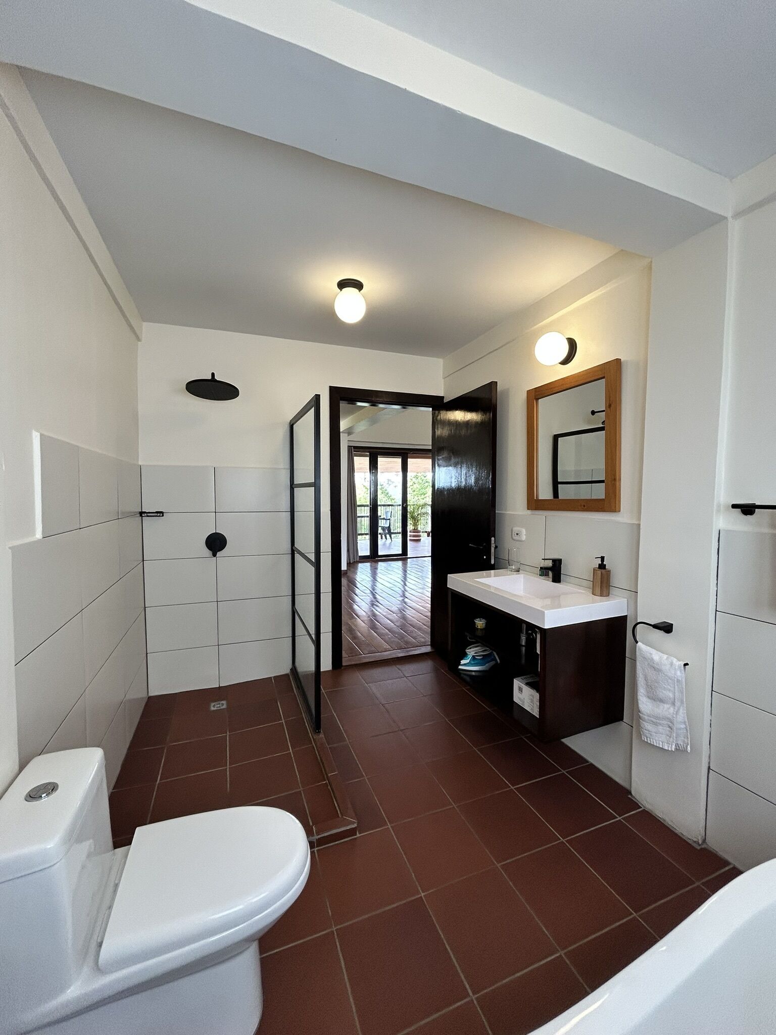 A modern bathroom with a toilet, sink, and shower. It features white tiles, a wooden door, and a view to an outdoor area.