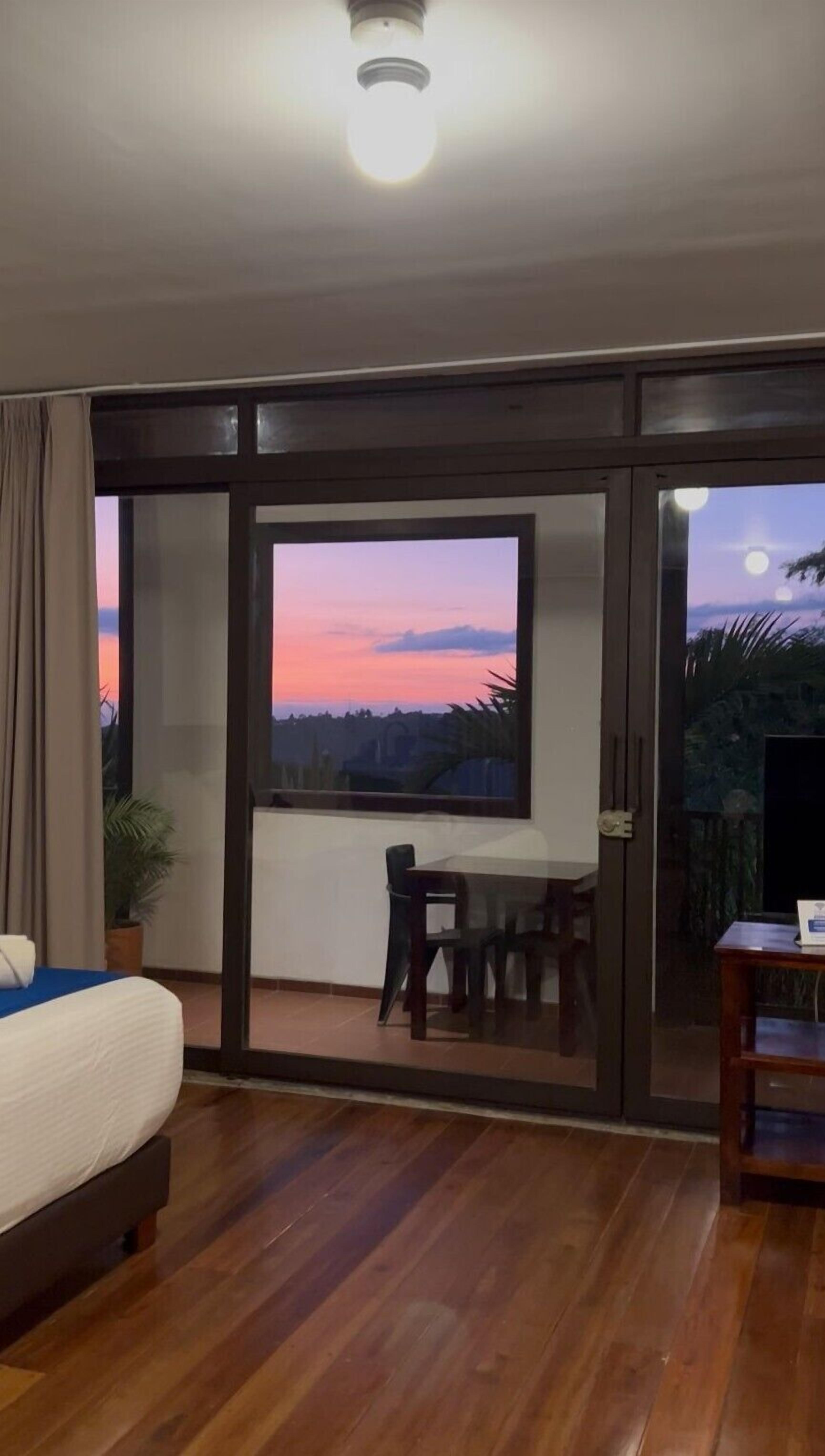 A bedroom with a bed, a desk, and a view of a sunset through a large window.