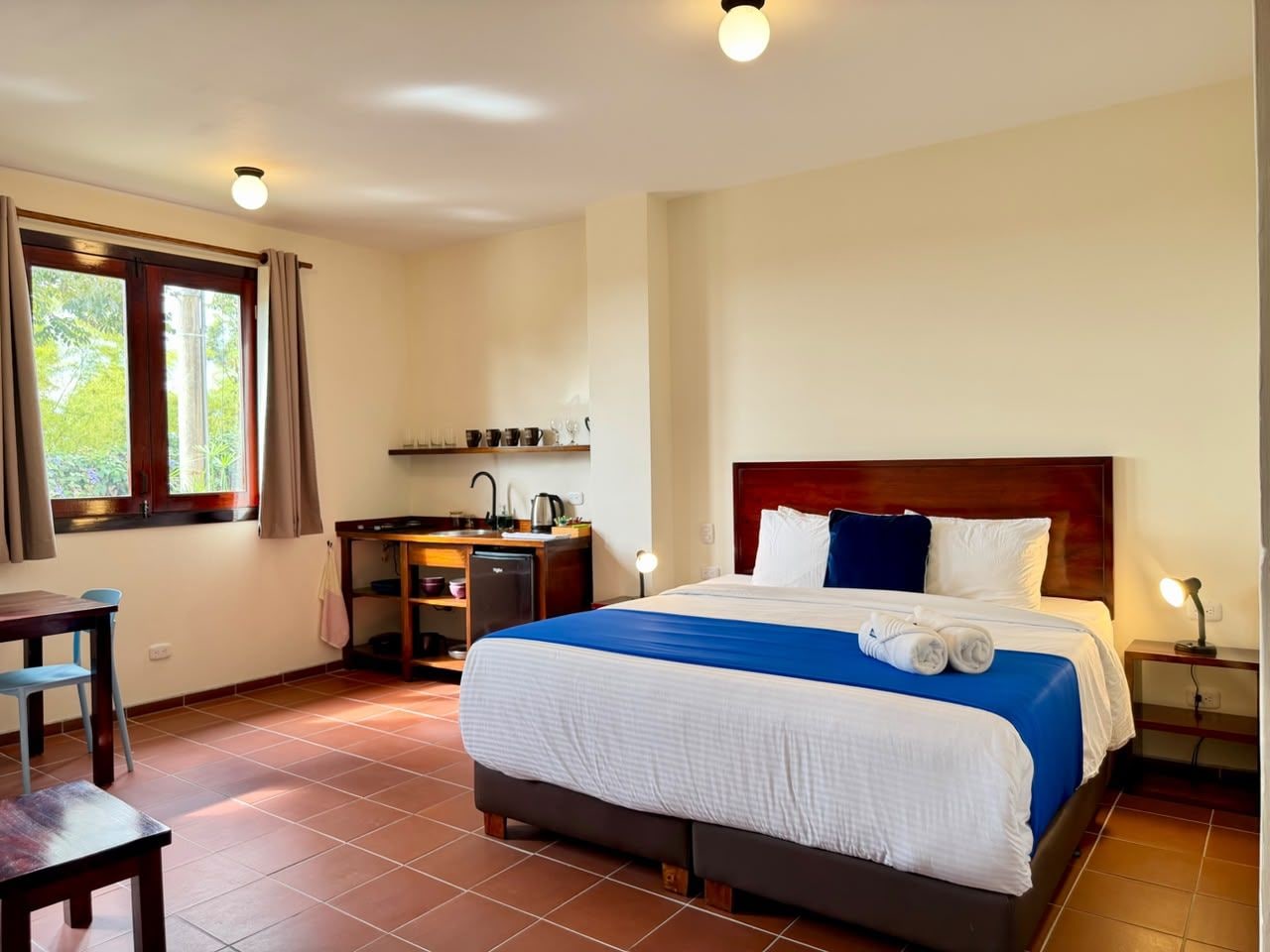 A hotel room with a bed, a desk, a small kitchenette, and a window with curtains.