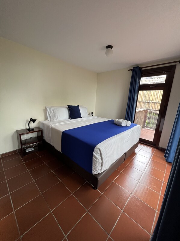 Un cuarto de hotel con cama, mesita de noche, ventana con cortinas y vista al balcón exterior.