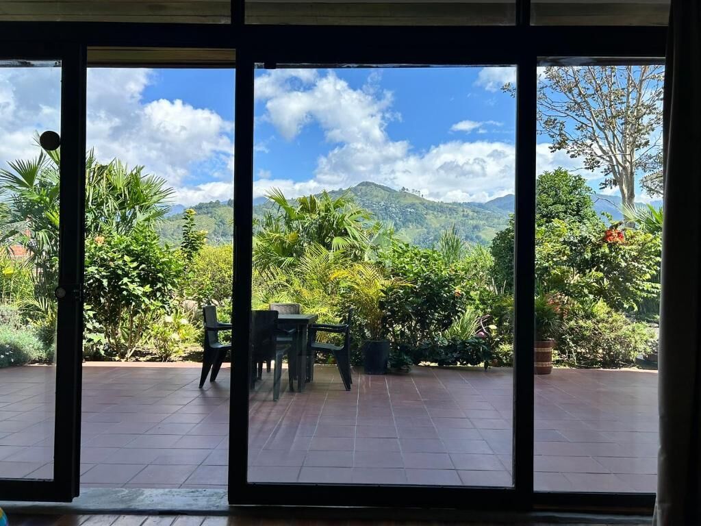 A view through a sliding glass door to an outdoor patio area with a table and chairs, surrounded by lush greenery and a mountain backdrop.