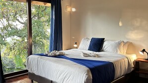 Un dormitorio con una cama grande, suelo de madera, techo con vigas a la vista, una ventana con cortinas y una mesita de noche con lámpara.