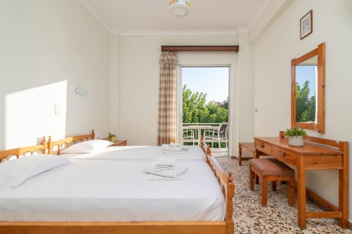 A hotel room with two beds, a wooden desk, a chair, and a balcony with a view of greenery.