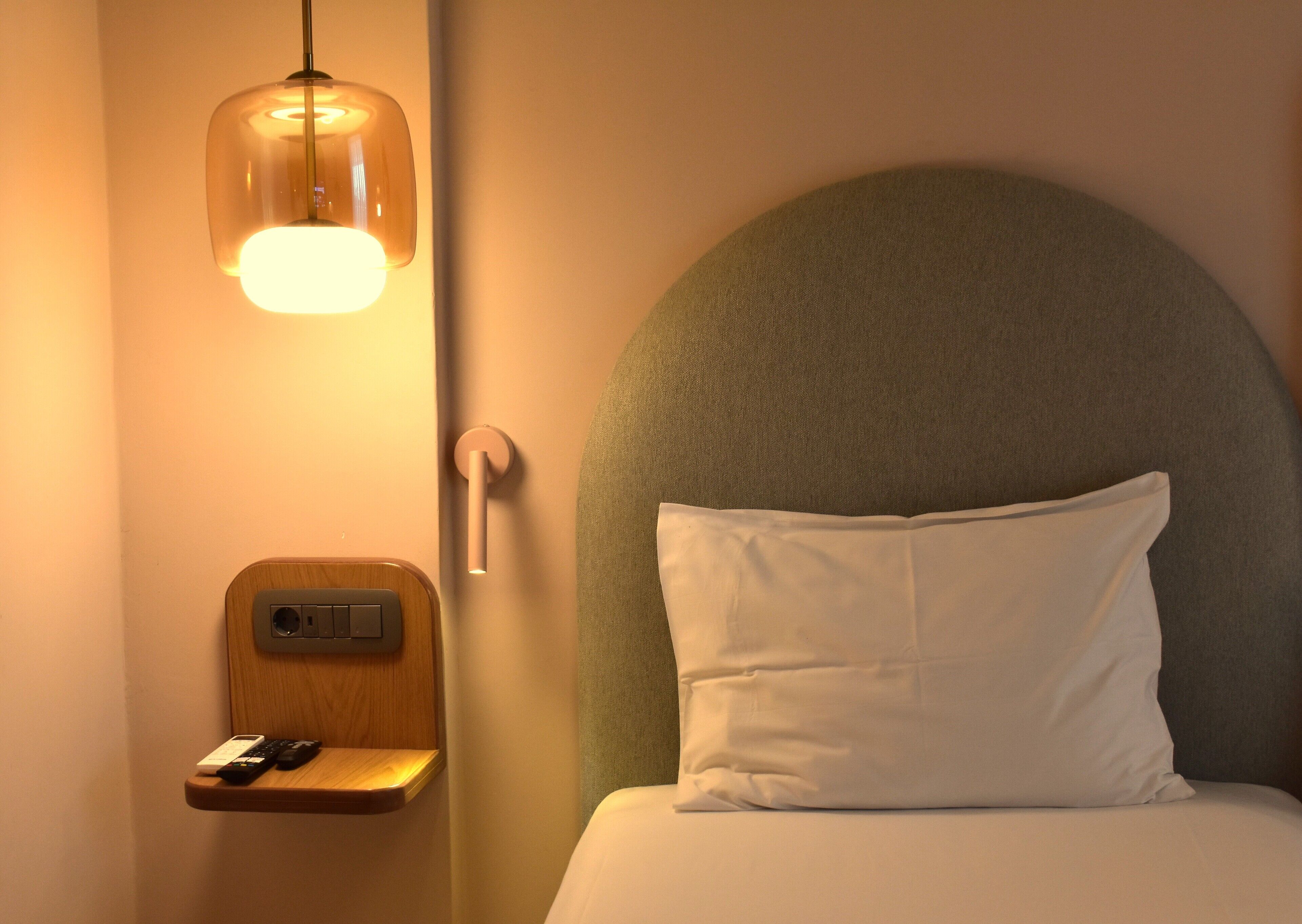 A neatly made bed with a white pillow and a headboard. A small wooden shelf holds a remote control and a phone. A wall-mounted light fixture casts a warm glow.
