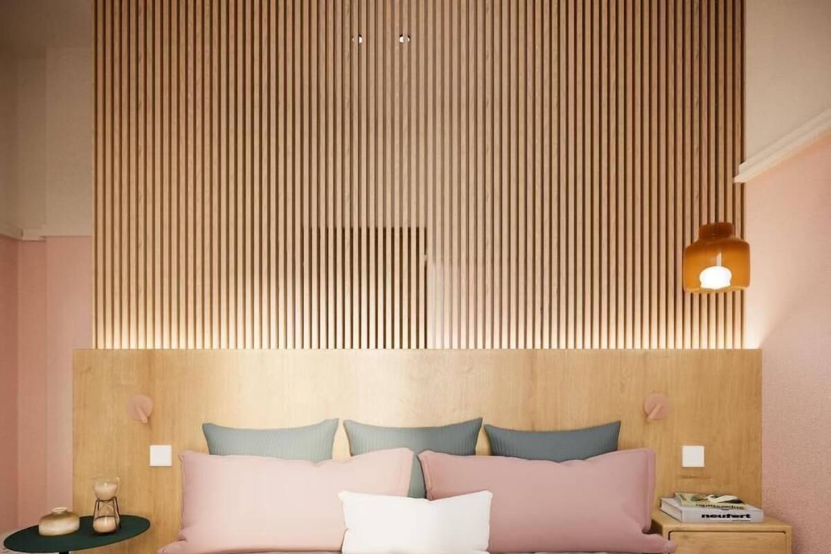 A bedroom with a wooden slatted headboard, a bed with pink bedding, a bedside table with a lamp, and a nightstand with a book.
