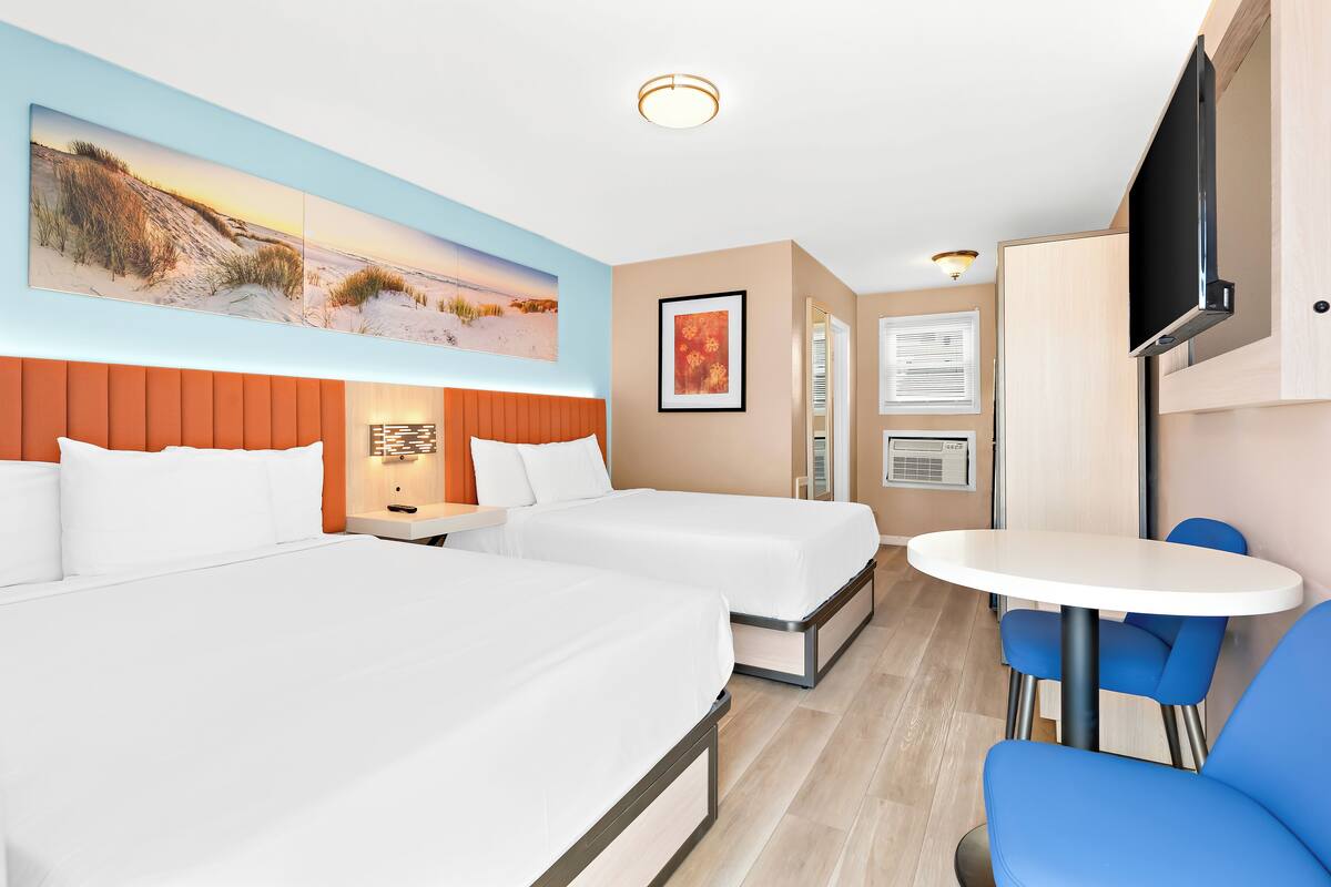 Ein Hotelzimmer mit zwei Betten, einem kleinen Tisch, blauen Stühlen, einem Fernseher und einem Bild an der Wand.