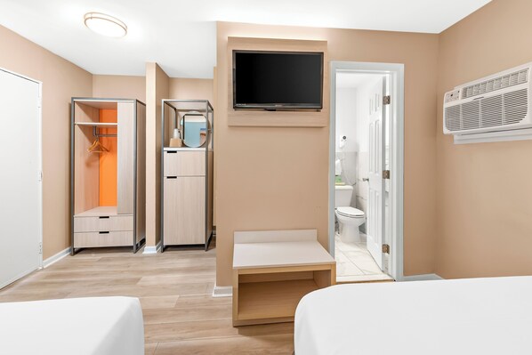 A hotel room with a bed, a TV mounted on the wall, a wardrobe, a mirror, and an air conditioning unit.