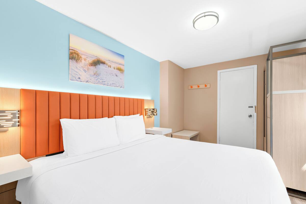 Ein modernes Hotelzimmer mit einem großen Bett, einem vertikal lamellenartigen Kopfteil, einem Gemälde mit Strandmotiv, einem Nachttisch mit Lampe und einem Kleiderschrank.