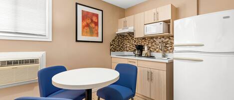A compact kitchen with a white refrigerator, a microwave, and a coffee maker on the countertop. There is a round table with blue chairs and a framed picture on the wall.