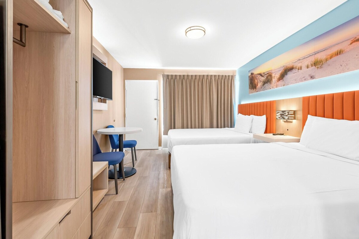 Quarto de hotel com duas camas, uma mesa com cadeiras, uma televisão e um quadro na parede.