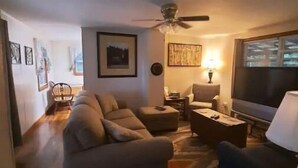 Un salon comprenant un canapé, des fauteuils, une table basse et un téléviseur. On y trouve également un ventilateur de plafond, une lampe et des tableaux encadrés accrochés aux murs.