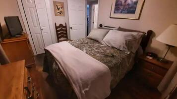 Een slaapkamer met een bed, een houten dressoir, een bureau met een computer, een lamp en een ingelijste foto aan de muur.