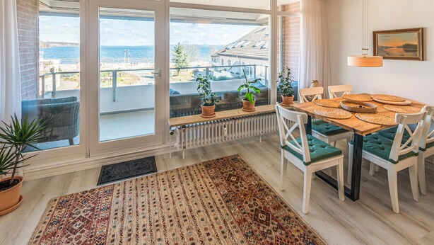 Zona pranzo con vista sul mare, tavolo apparecchiato con sottopiatti intrecciati e tappeto decorativo sul pavimento.