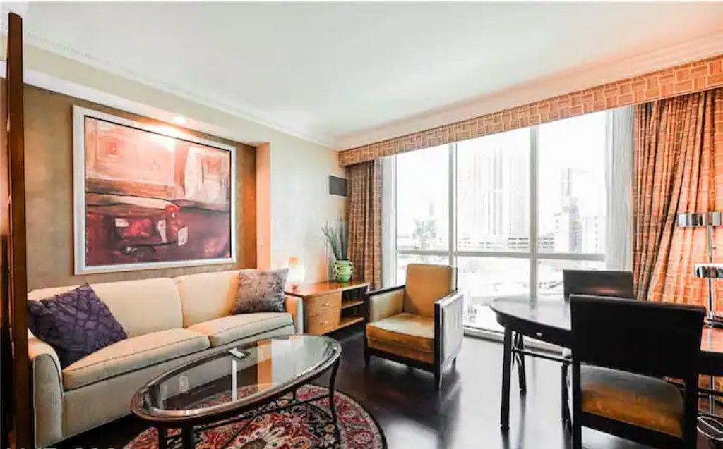 A modern living room with a sofa, coffee table, and chairs. There is a large window with curtains, a painting on the wall, and a city view.