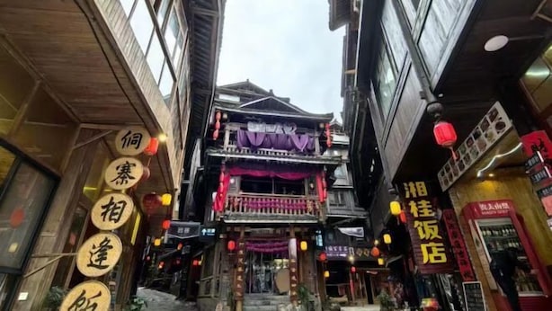 Une rue étroite bordée de bâtiments traditionnels en bois, une boutique avec une enseigne en caractères chinois et un immeuble surmonté d’un auvent violet.