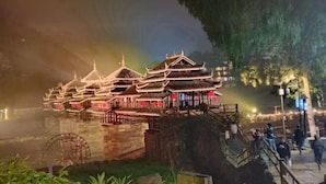 Un bâtiment traditionnel à plusieurs niveaux, doté d’une roue à eau, entouré de verdure et d’un mur de pierre.