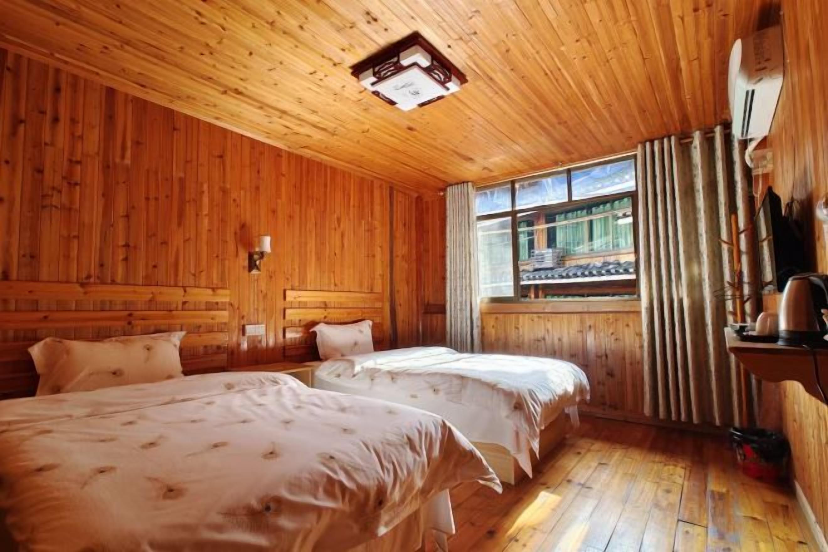 A wooden cabin room with two beds, a window with curtains, a lamp, and a wooden ceiling.