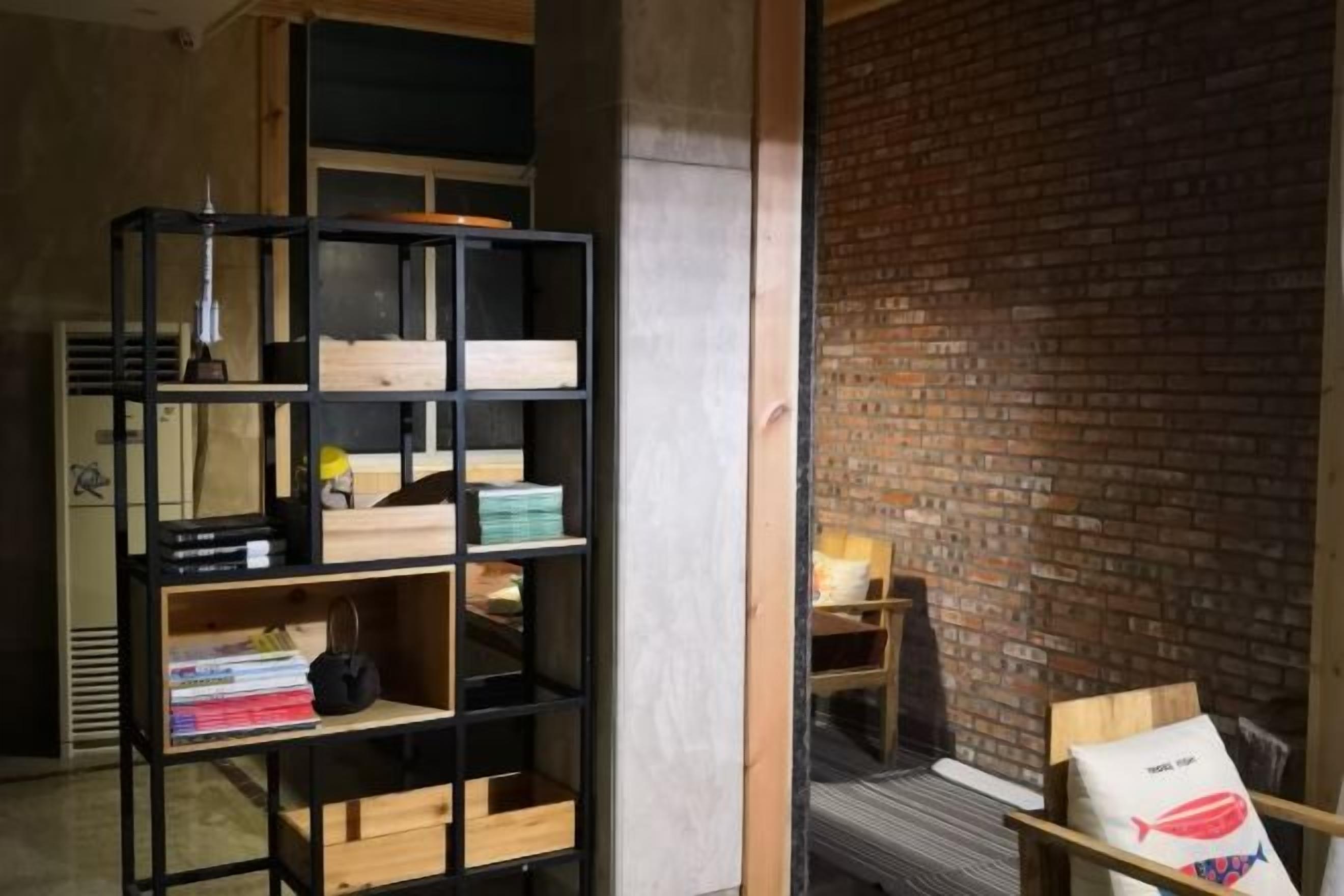 A room with a brick wall, a black shelving unit with various items, and a wooden chair.