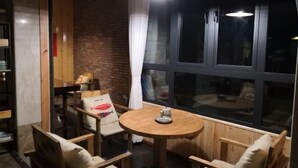 Un rincón acogedor con una mesa redonda de madera y sillas acolchadas, una pared de ladrillo y un amplio ventanal con cortinas.
