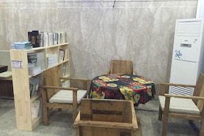 Une pièce comprenant une chaise en bois, une petite table recouverte d’une nappe colorée, une bibliothèque et un réfrigérateur blanc.