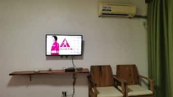 Una habitación con televisión de montaje, un estante de madera, dos sillas de madera y aire acondicionado.