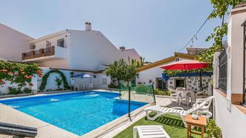 Une piscine entourée d’un patio aménagé avec des chaises longues et une table, un espace de détente couvert avec un parasol rouge, et un bâtiment à la façade blanche ornée de carreaux décoratifs.