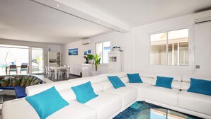 Sala moderna com sofá branco, almofadas azuis, mesa de centro de vidro e área de jantar com cadeiras transparentes.