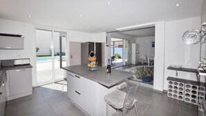Cozinha moderna com ilha central, área de jantar com mesa de vidro e vista para uma piscina.