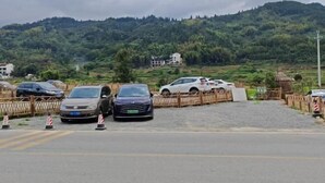 Un estacionamiento con varios coches, una valla de madera y un paisaje montañoso al fondo.