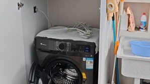 Un lavadero pequeño y funcional con lavadora de carga frontal, tendedero y un estante con productos de limpieza.