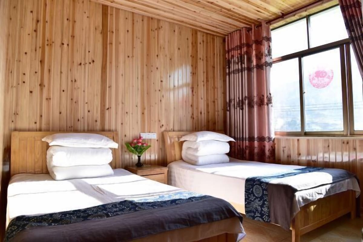 A wooden cabin room with two beds, a window with curtains, and a small table with a vase of flowers.