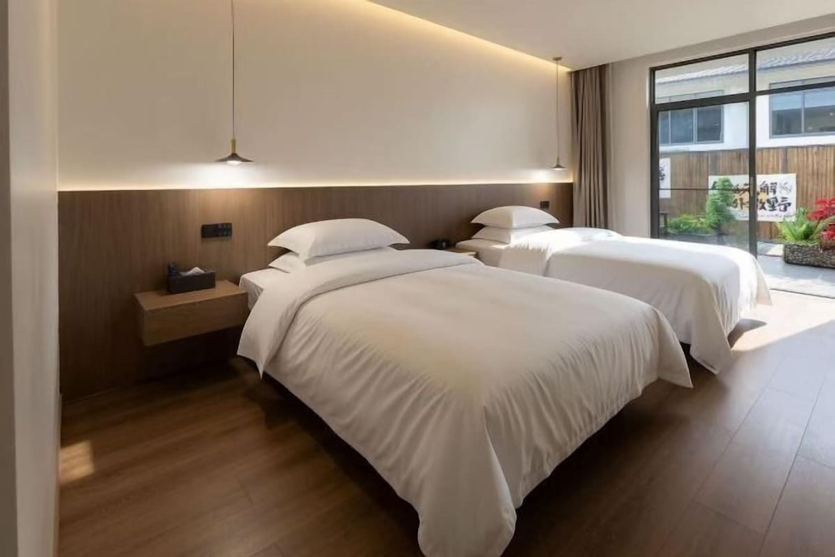 A modern bedroom with two beds, a bedside table, and a large window.