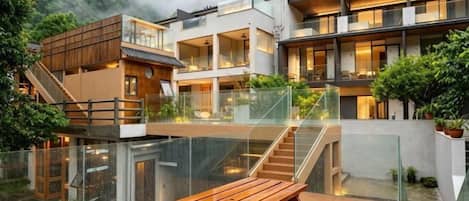 Un edificio moderno de varios pisos con barandas de vidrio, bancos de madera al aire libre y una escalera que sube a niveles superiores.