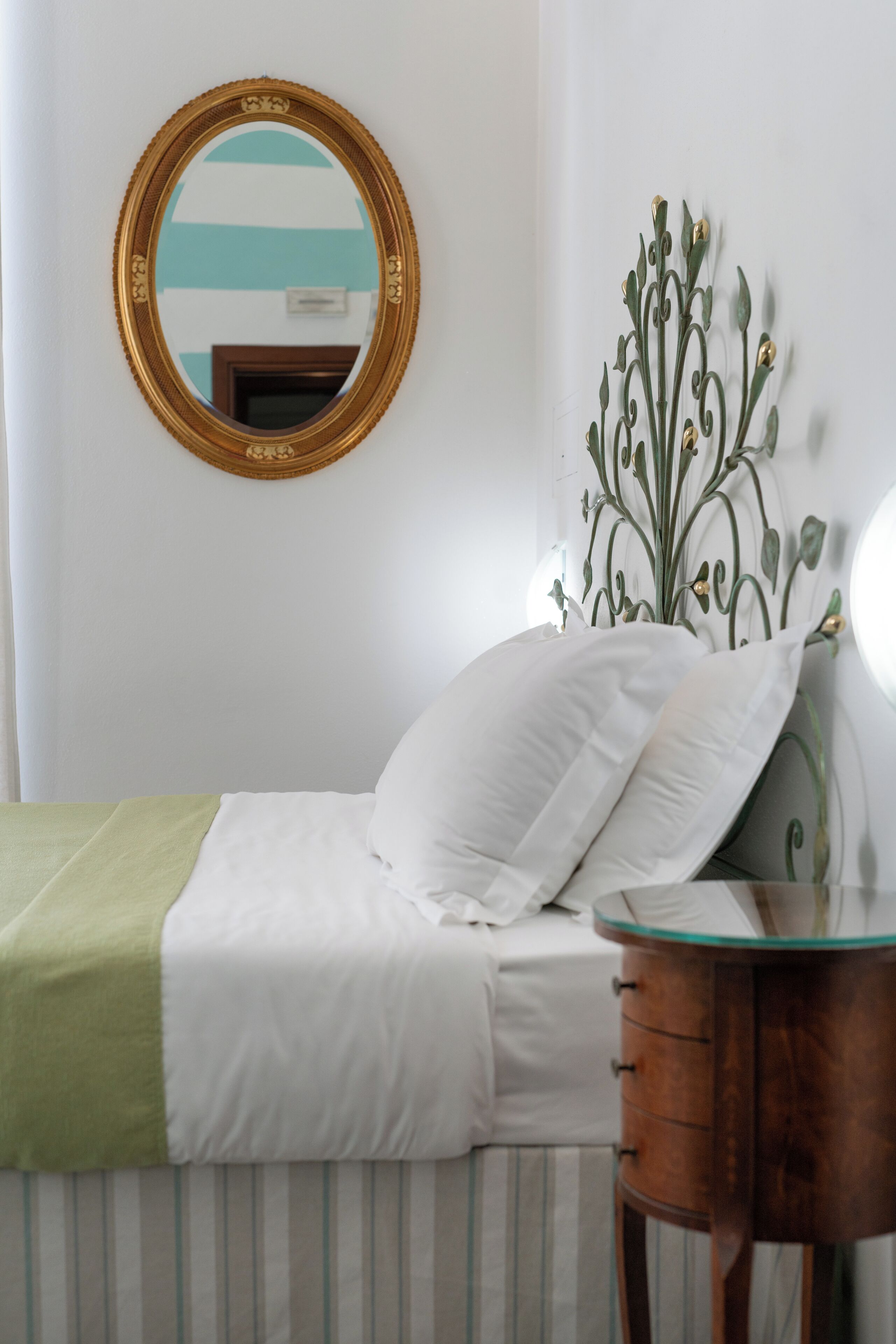 A neatly made bed with white bedding and a green throw, a wooden bedside table, a wall-mounted mirror, and a decorative metal plant.
