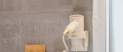 A bathroom with a hairdryer mounted on the wall, a glass shelf with toiletries, and a mirror with ornate detailing.