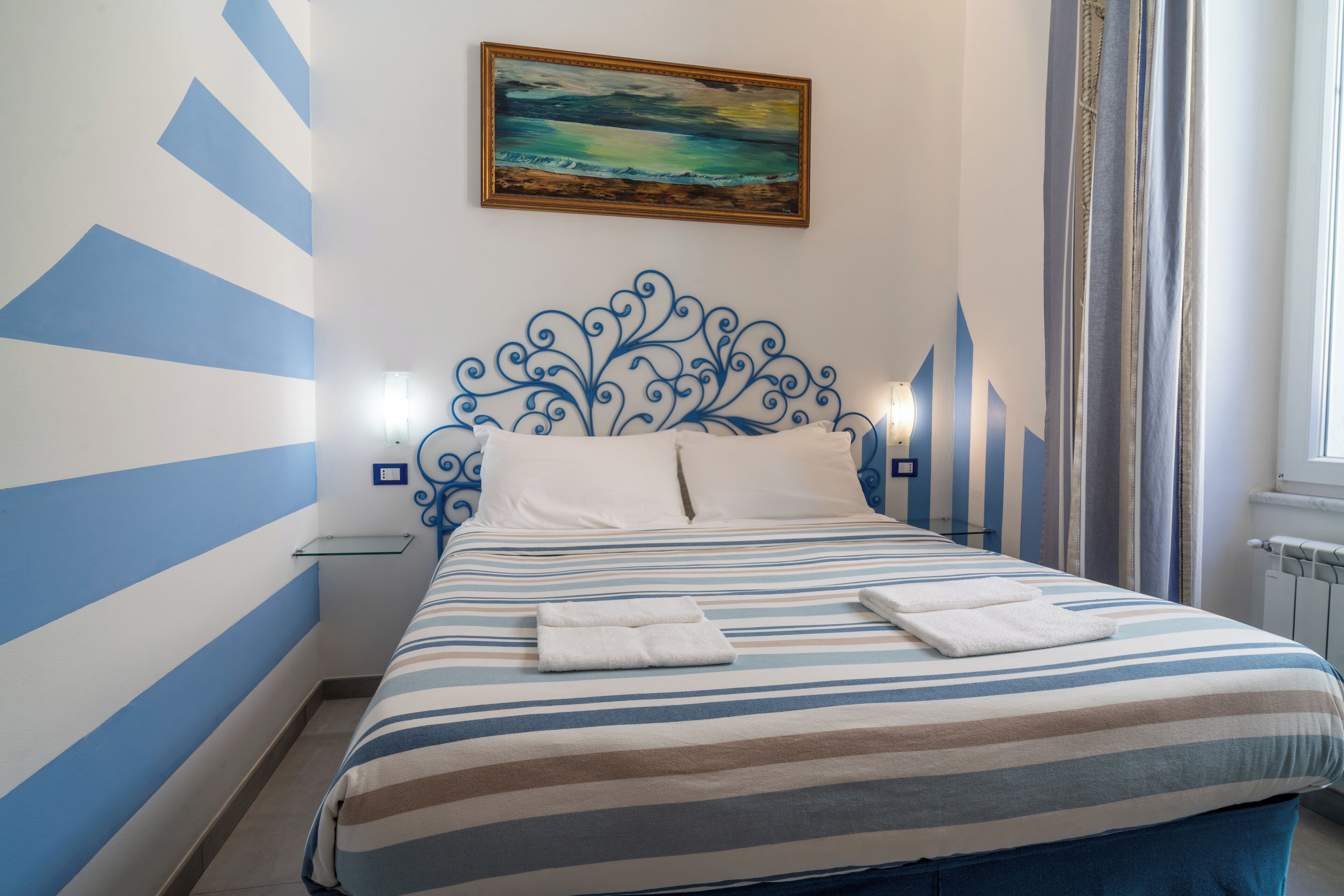Una habitación de hotel con ropa de cama a rayas, un cabecero decorativo, un cuadro en la pared y una ventana con cortinas.