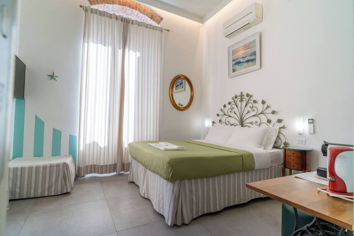 Une chambre d’hôtel comprenant un lit, un bureau, une chaise, une petite table, un miroir et une fenêtre avec des rideaux.
