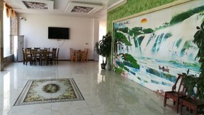 A spacious lobby with a large mural of a waterfall, a flat-screen TV mounted on the wall, a dining area with wooden chairs, and a polished tiled floor.