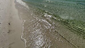 A beach with clear water, a sandy shore, and a bright sun in the sky.
