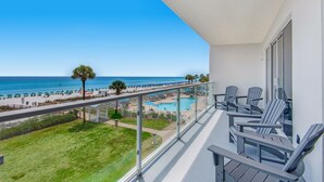 A balcony with a view of a pool and beach, and several Adirondack chairs.