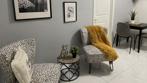 A room with a grey wall, a white door, two leopard print chairs, a small round table with a clock and a plant, and a shelf with decorative items.